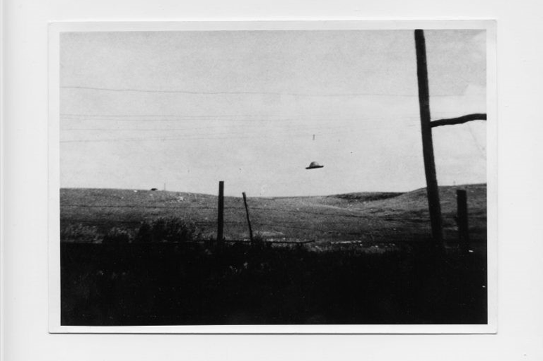 Large field, fence posts in the foreground, a small disc-like object resembling an unidentified flying object near centre-frame hovering in the sky in the distance.