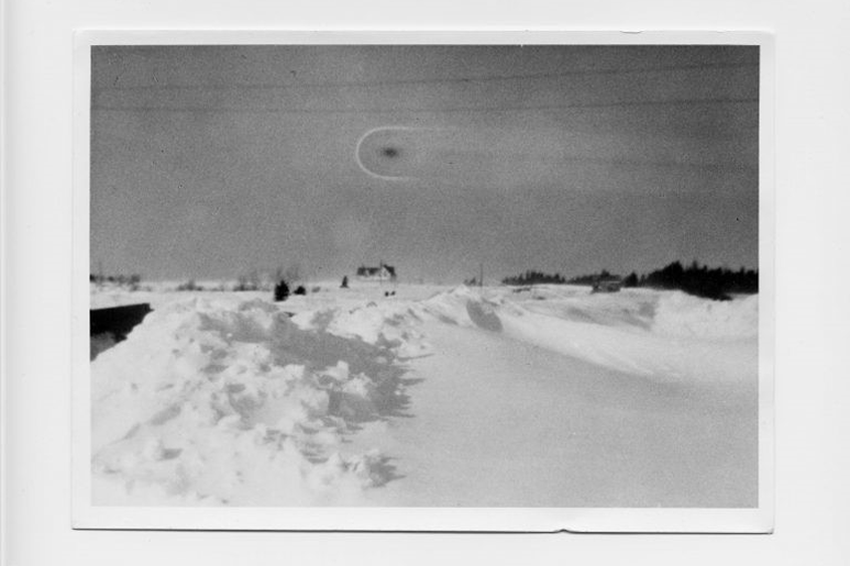 Large open snowy field, a farmhouse visible far in the distance, in the sky a circular black smudge that could be taken to be an unidentified flying object.