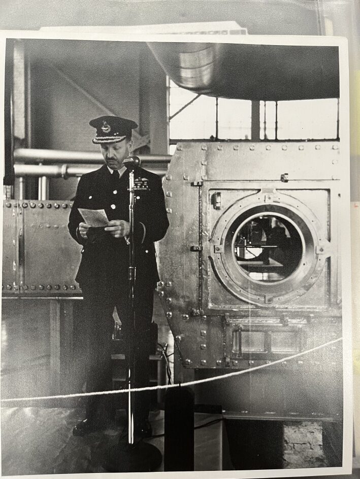 A Royal Canadian Air Force officer speaking before a wind tunnel apparatus.