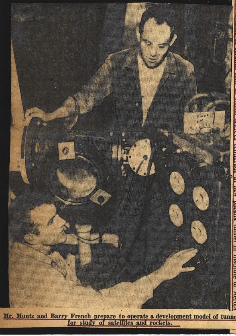 Two engineers (Phillip Muntz and Barry French) in work clothes make adjustments to the dials of a large piece of lab equipment.