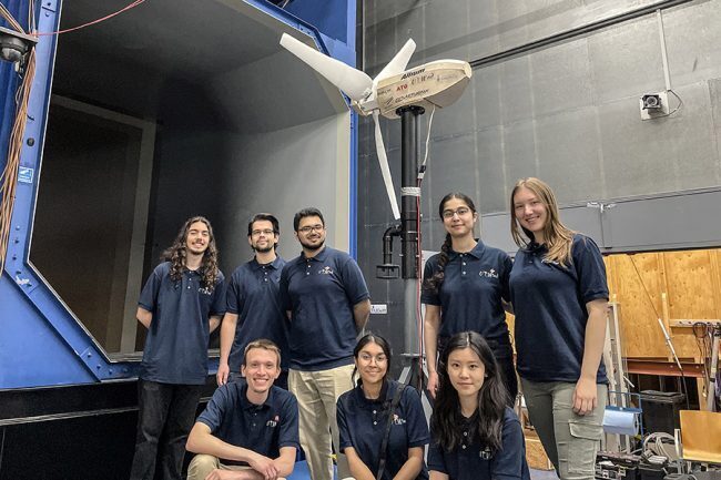 The UTWind team of eight people next to their winning prototype turbine at the Open Jet Facility wind tunnel at Delft University of Technology