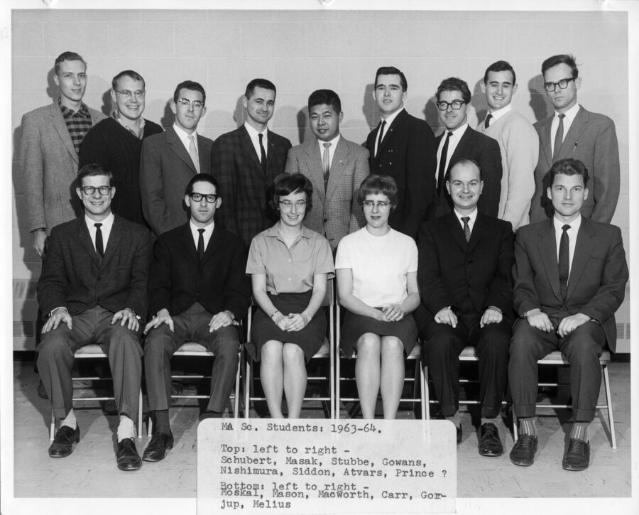 A group of MA.Sc. students pose for a photo inside, 1963-64. Two women are present amongst the male students, Jean MacWorth and Donna Carr.