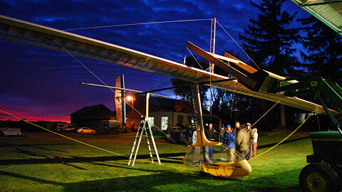 A large ornithopter rests on a nighttime field, a team of engineers grouped beside it.