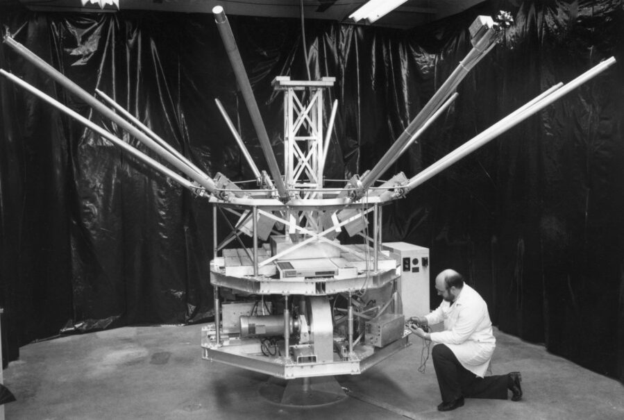 An engineer in a lab coat kneels before the satellite-like structure of Daisy, long struts jutting up and out from its central hub.