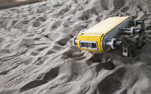 Six-legged yellow metal robot crawls across sandy terrain in the University of Toronto Institute for Aerospace Studies MARS Dome.