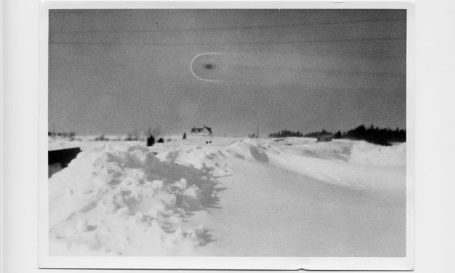 Large open snowy field, a farmhouse visible far in the distance, in the sky a circular black smudge that could be taken to be an unidentified flying object.