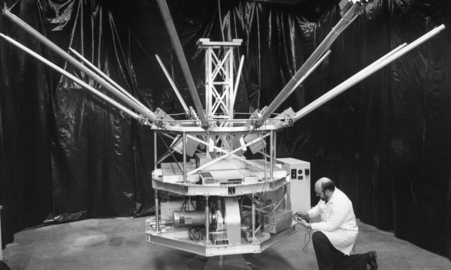 An engineer in a lab coat kneels before the satellite-like structure of Daisy, long struts jutting up and out from its central hub.
