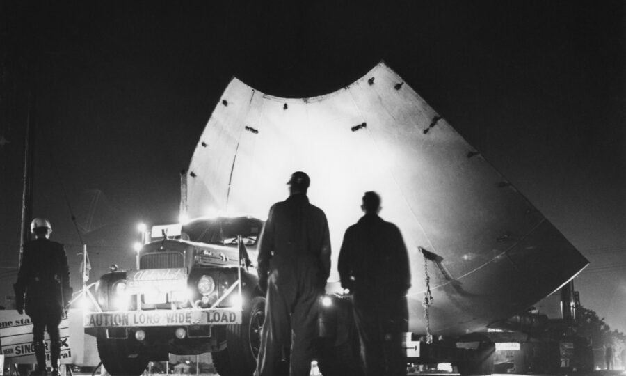 At night, two workmen and a police officer observe a transport truck loaded with a section of the giant metal sphere being transported to the new UTIAS location.